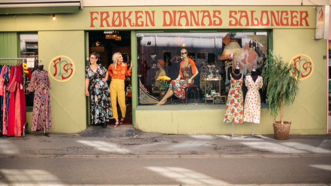 To personer står utenfor «Frøken Dianas Salonger», en sjarmerende vintage klesbutikk. Mannequins og fargerike kjoler fyller vinduet i denne herlige bruktbutikken, og inviterer forbipasserende til å utforske retroskatter innenfor.
