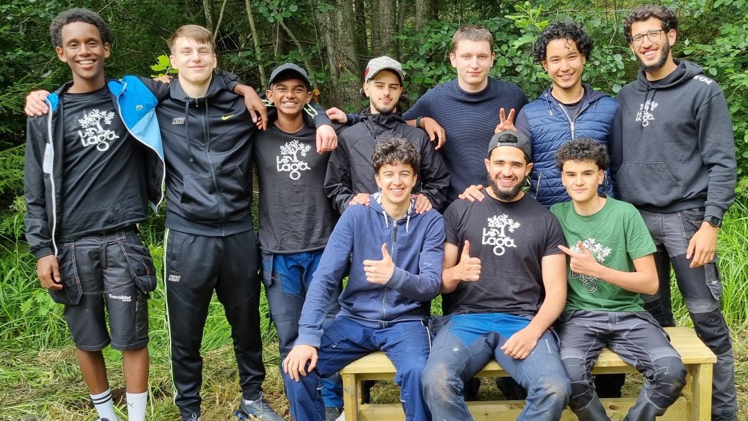 En gruppe på ti personer, noen stående og noen sitter på en benk, uformelt kledd blant frodig grønt løvverk og trær som bidrar til det rolige bakteppet.