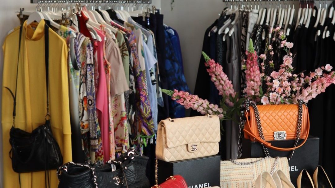 Klær og håndvesker er kunstferdig utstilt i en sjarmerende butikk som minner om trendy bruktbutikker. Fargerike plagg henger på stativer ved siden av hyller med designervesker og sko, mens rosa blomster fremhever arrangementet.