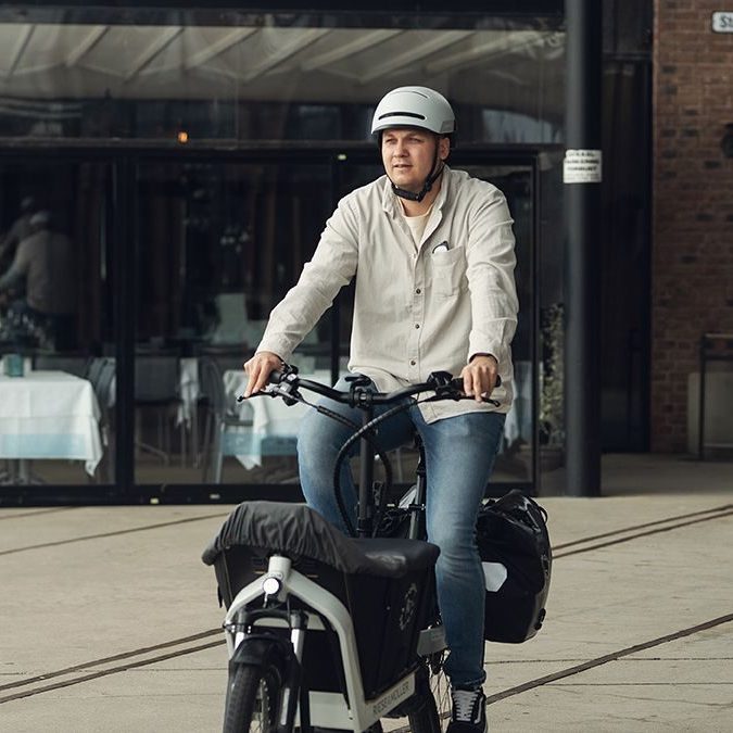 En person med hjelm sykler på en lastesykkel, eller elektrisk lastesykkel, gjennom et asfaltert byområde med elegante glassbygninger i bakgrunnen.