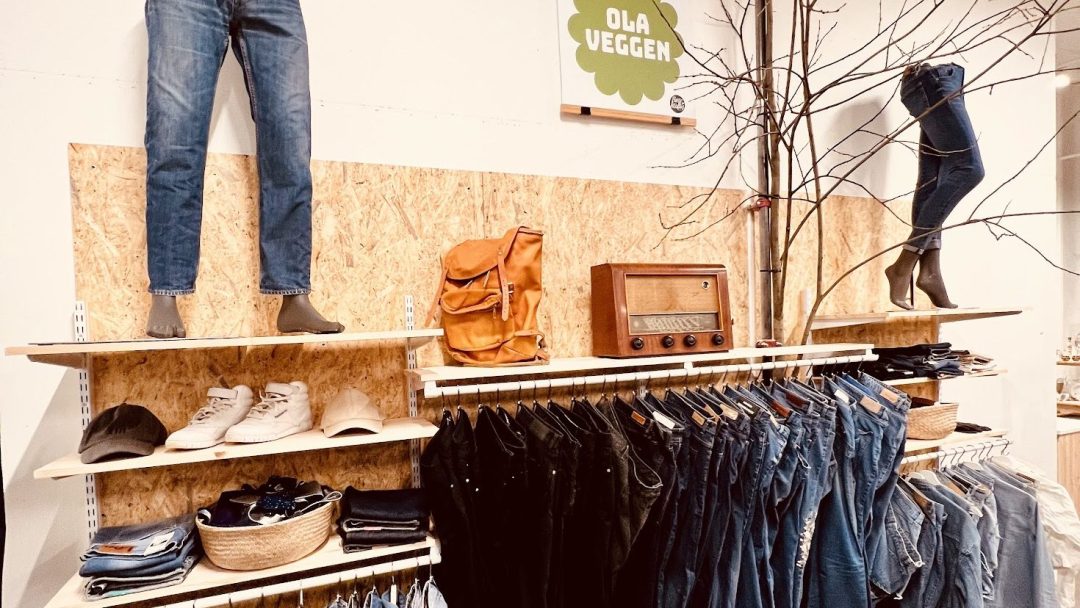 Klesbutikken viser kunstferdig jeans, sko, skjorter, en oransje ryggsekk og en vintage radio i trehyller. På et skilt står det OLA VEGGEN, som fremkaller sjarmen til bruktbutikker.