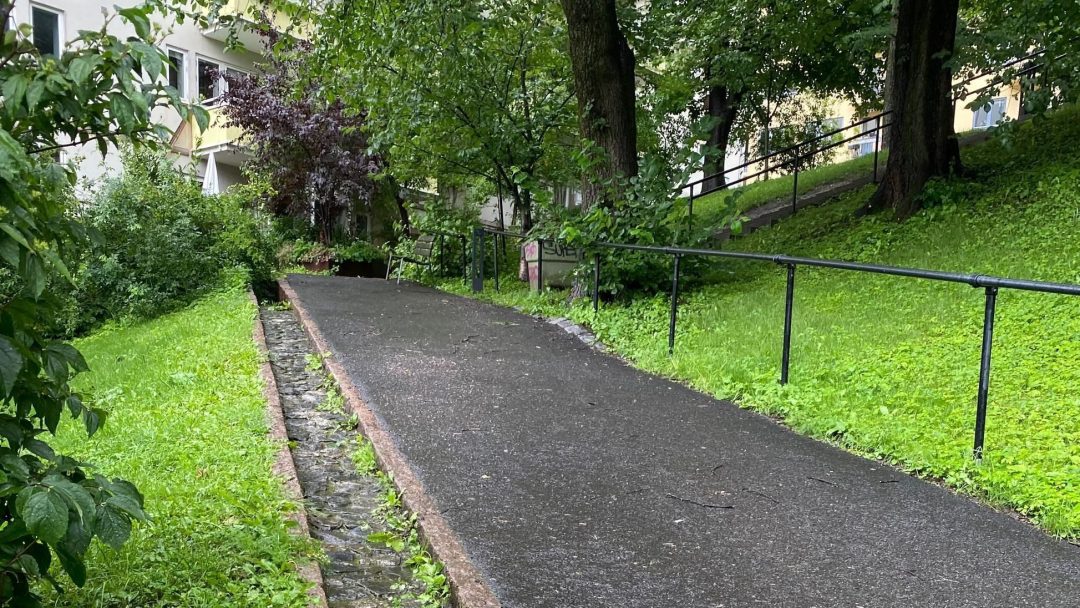 En asfaltert sti fører oppover, flankert av frodig gress og trær. Et rekkverk langs høyre side, med en bygning delvis synlig til venstre under en overskyet himmel. Denne rolige scenen ligger stille innenfor de større rammene av klimabudsjettets hensyn til bærekraftig byplanlegging.