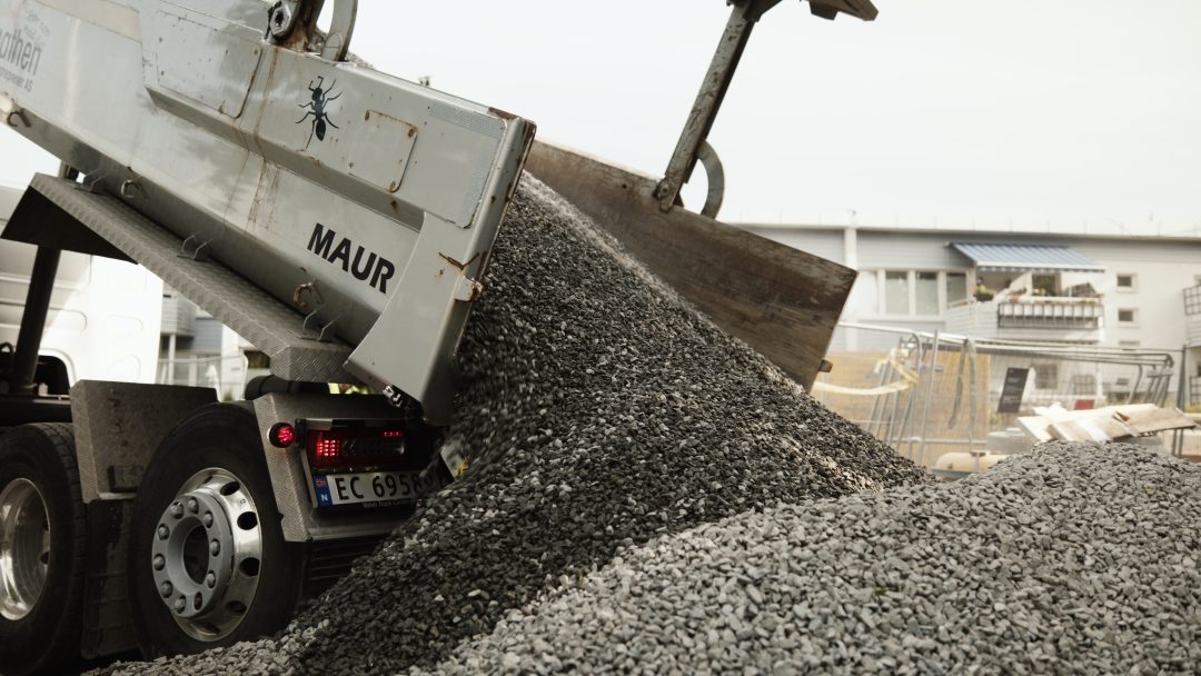 En lastebil losser grus på en byggeplass, og bidrar til infrastrukturutviklingen innenfor rammen av klimabudsjettet. En haug med grus bygger seg opp under den høye lastebilen som en del av dette miljøbevisste prosjektet.