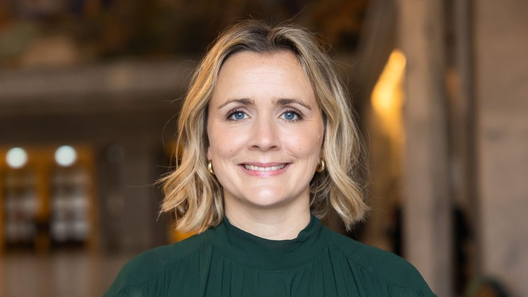 En person med skulderlangt blondt hår, iført en grønn topp, smiler mykt mot en uskarp innendørs bakgrunn. Hennes fredelige oppførsel er beslektet med den stille effektiviteten til hurtigladere for tunge kjøretøy.