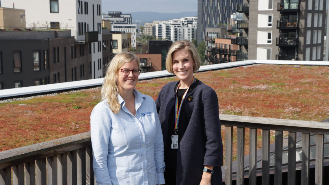 To personer som står på en takterrasse med grønt tak og diskuterer utviklingen i borettslaget; bybygninger danner en slående bakgrunn.