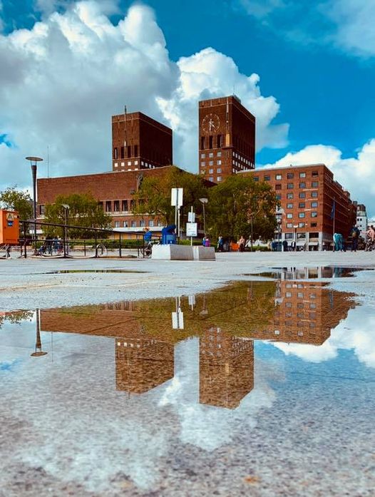 Mursteinsbygning med to tårn, med en klokke, reflekteres i en sølepytt. Trær og mennesker er synlige under en delvis overskyet himmel.