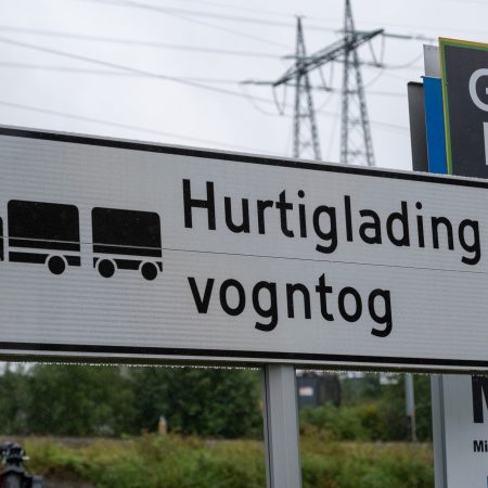 Skilt for Hurtiglading vogntog, som indikerer hurtigladestasjoner for lastebiler, med et lastebilsymbol. Kraftledninger og grønne omgivelser rammer inn bakgrunnen, og fremhever tilgjengeligheten av hurtigladere for tunge kjøretøy.
