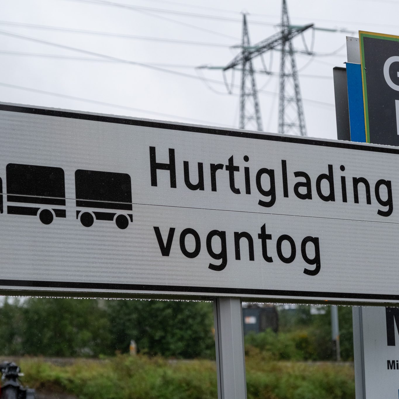 Skilt for Hurtiglading vogntog, som indikerer hurtigladestasjoner for lastebiler, med et lastebilsymbol. Kraftledninger og grønne omgivelser rammer inn bakgrunnen, og fremhever tilgjengeligheten av hurtigladere for tunge kjøretøy.