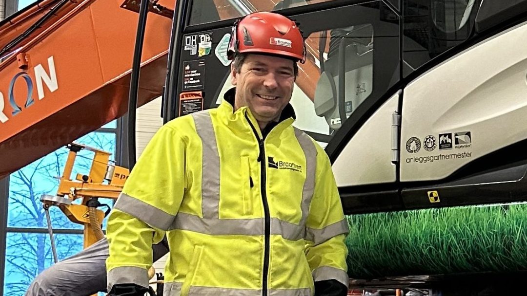 En person i fluorescerende gul jakke og rød hjelme står foran en elektrisk lastebil og tunge maskiner inne i en bygning.