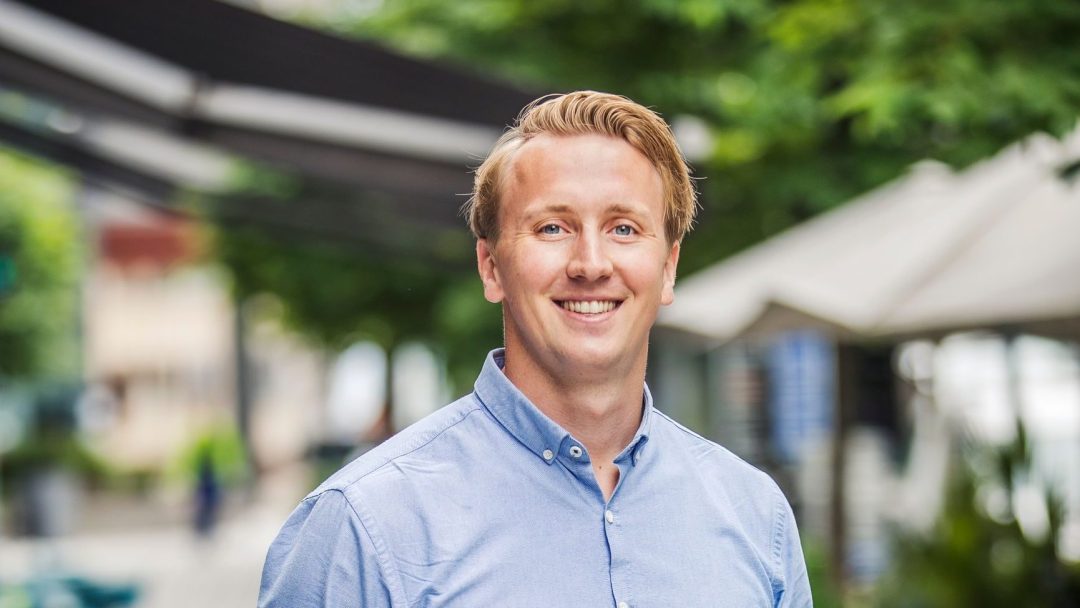 En person med blondt hår smiler lyst, iført en blå skjorte, og står utendørs i en livlig gate. Scenen kan skilte med trær og markiser i bakgrunnen, og fanger essensen av et energifleksibilitetsmerket yrende potensial.