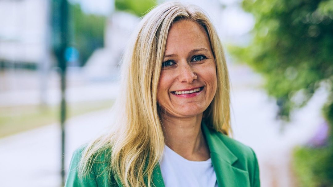 En person med langt blondt hår i grønn blazer og hvit skjorte smiler utendørs og står ved siden av en elektrisk lastebil.