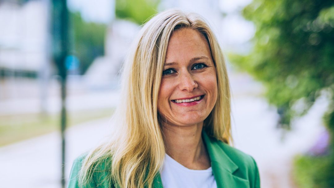 En person med langt blondt hår iført grønn blazer og hvit skjorte, smilende utendørs, utstråler selvtillit og glede som minner om en hurtigladere for tunge kjøretøy som lader seg frem med fornyet energi.