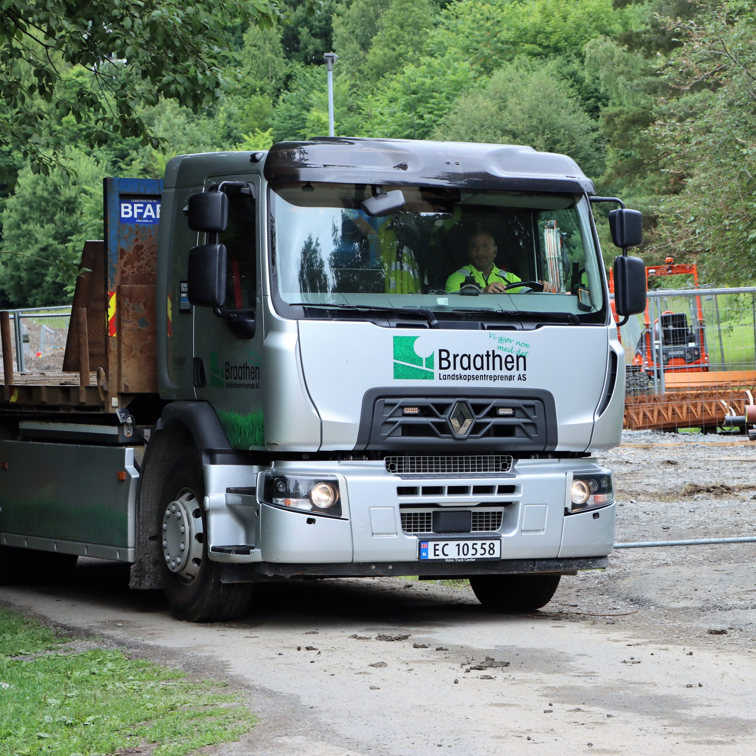 En grå elektrisk lastebil med Braathen-logoen frakter materialer på en grussti nær et inngjerdet område, omgitt av grøntområder.