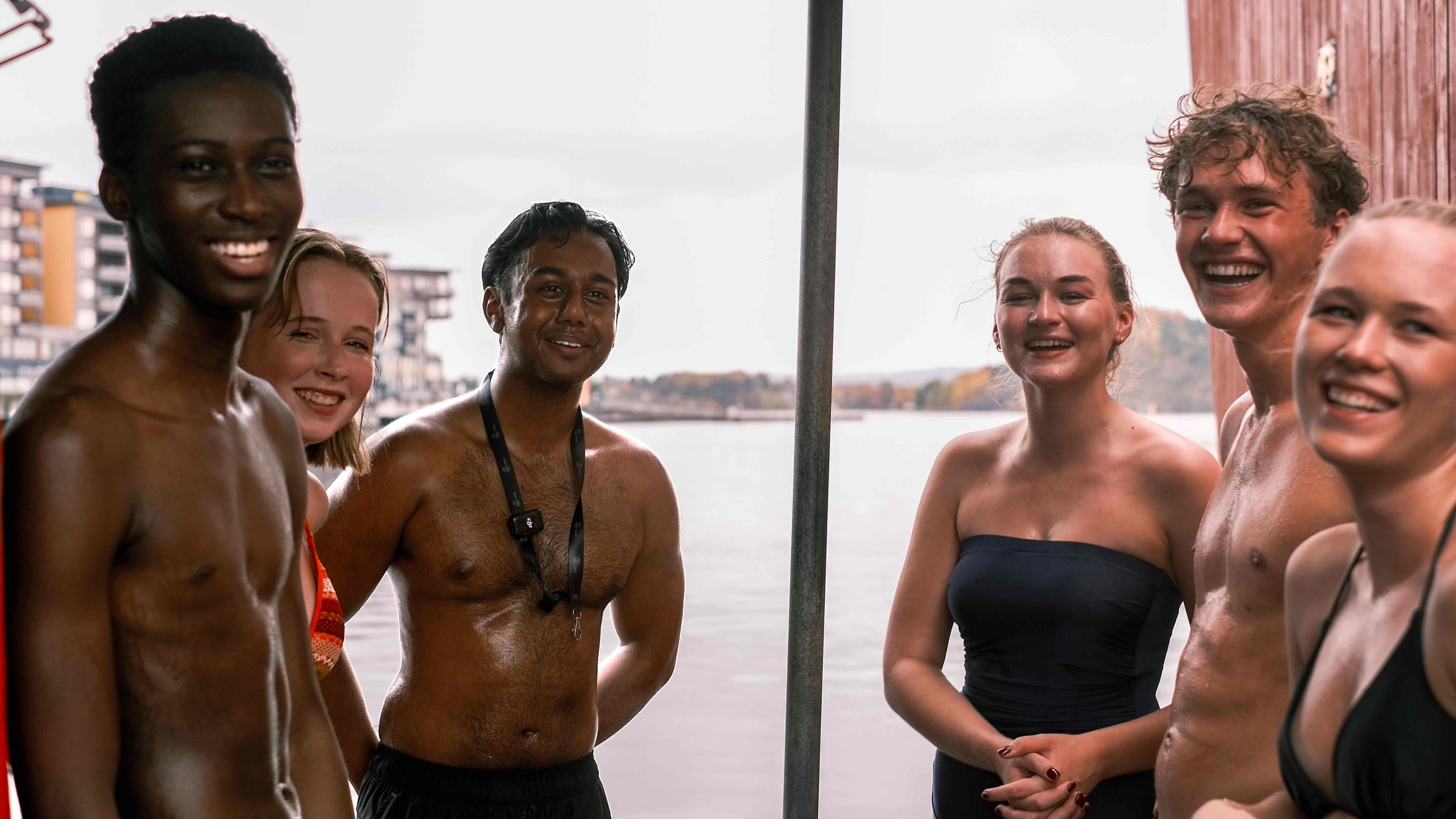 En gruppe på seks personer i badedrakter står sammen nær en rolig vannmasse og smiler varmt til kameraet mot et bakteppe av fjerne bygninger, som om de nettopp har hatt en forfriskende dukkert i badstuen.