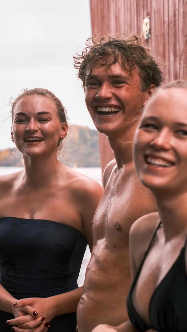 Tre personer i badedrakter smiler mens de står utendørs i nærheten av en badstu i tre, med uskarpt natur i bakgrunnen.