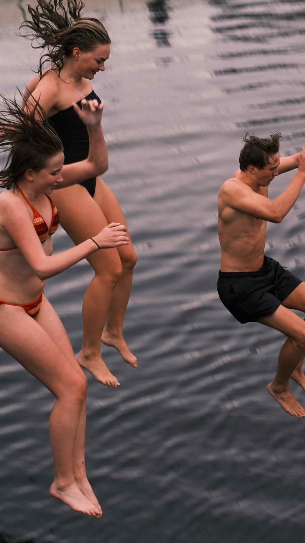 Tre personer i badetøy hopper glade fra kanten og dykker ned i badstuens forfriskende vann.