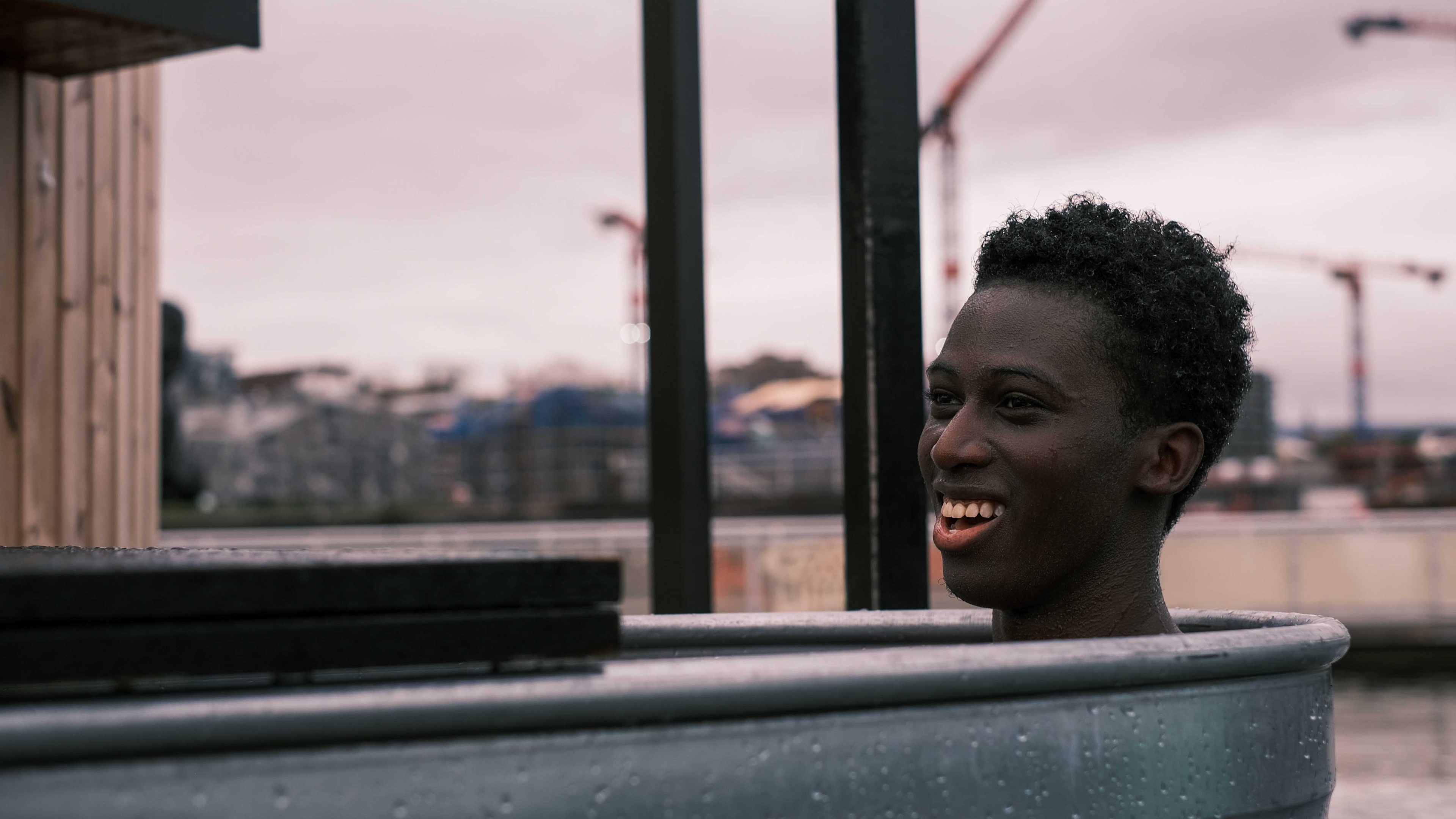 En person smiler mens han står i en badstu, en utendørs vanntank i metall, med industrielle strukturer i bakgrunnen.