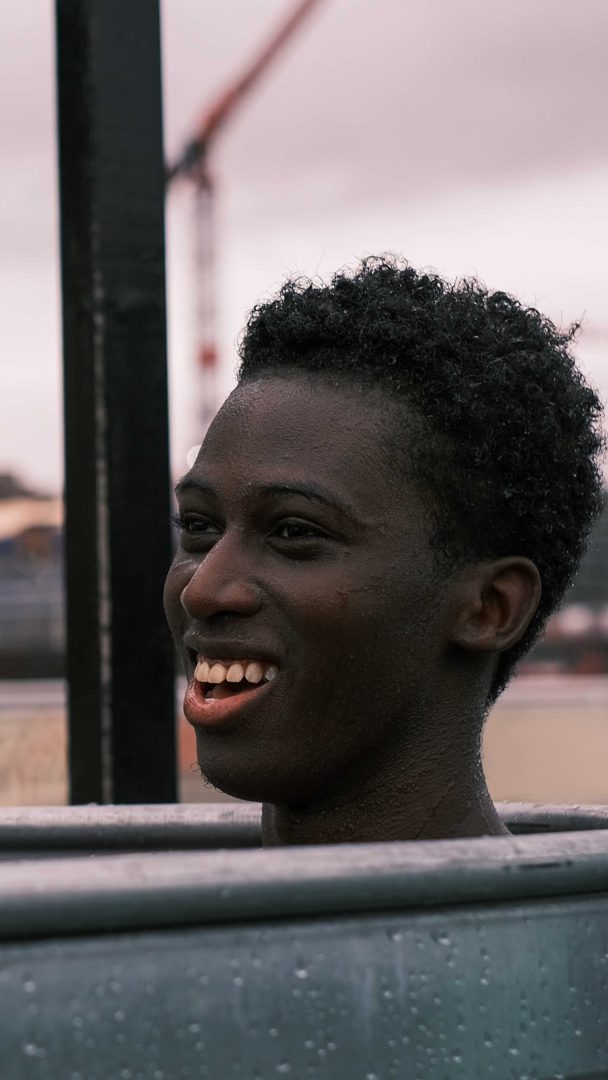 En person med kort, krøllete hår smiler utendørs, delvis nedsenket i et sjarmerende badstubadekar. Bakgrunnen har en uskarp kran satt mot en overskyet himmel.