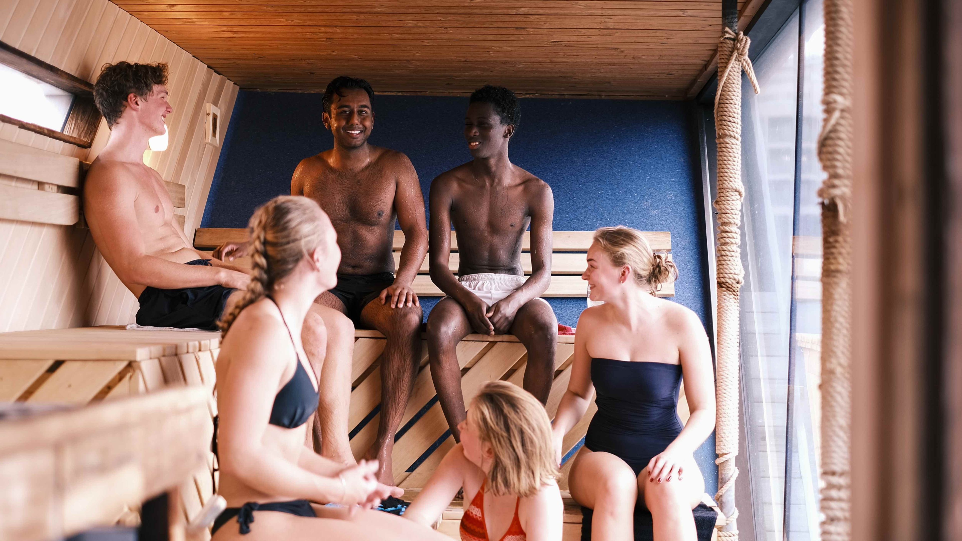 Seks personer i badetøy sitter og prater inne i en badstu i tre, med benker langs veggene og et vindu som gir lys.