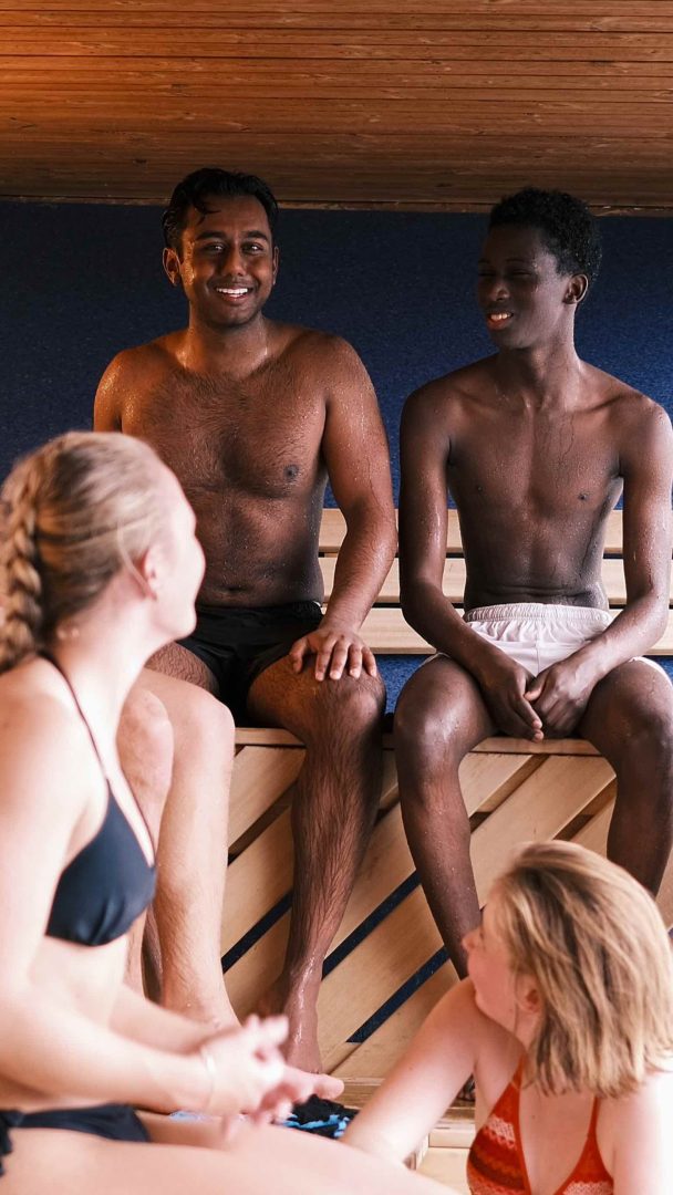 Fire personer sitter på trebenker i en koselig badstu, iført badedrakter og snakker.