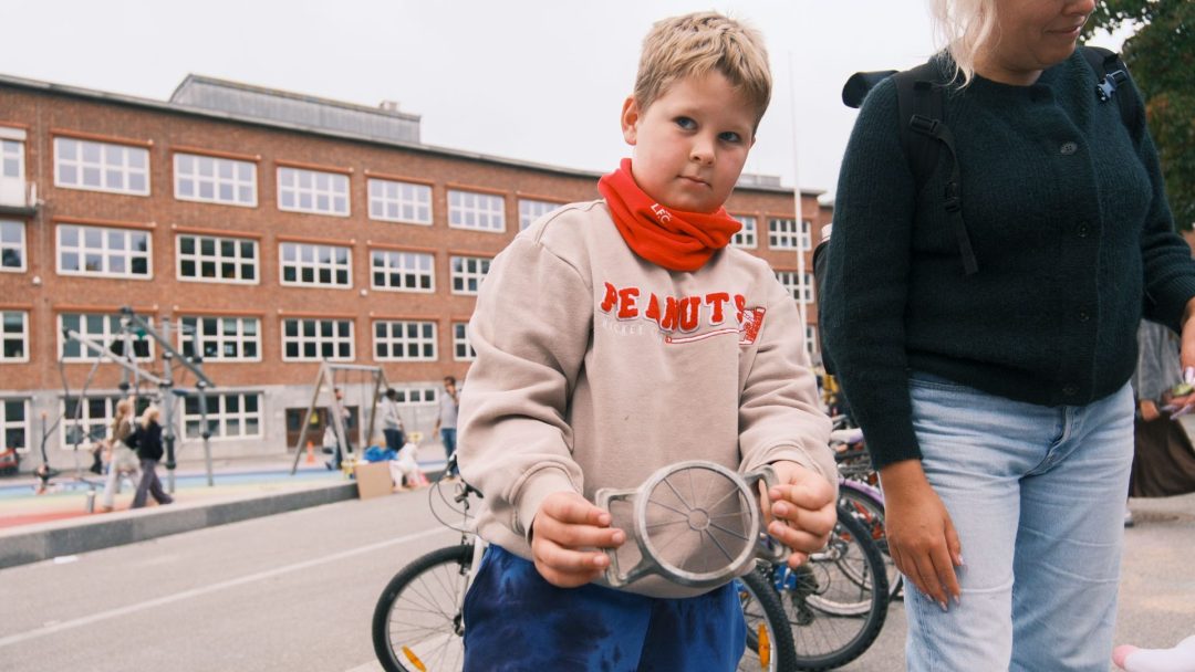 Et barn holder en rund gjenstand foran en skolebygning, med sykler og en lekeplass i bakgrunnen, som om de ser på skatter funnet på en lokal loppis. I nærheten står en annen person og observerer åstedet.