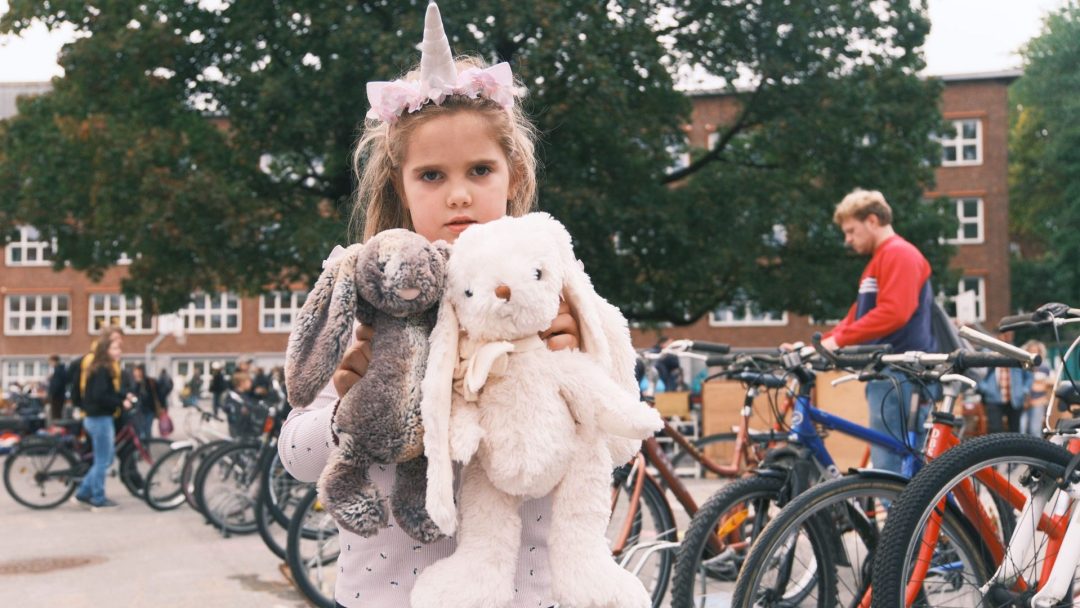Ung jente med pannebånd som ligner et enhjørningshorn holder to utstoppede kaniner, som minner om et sjarmerende loppisfunn, foran et sykkelstativ og trær, med mennesker og en bygning i bakgrunnen.