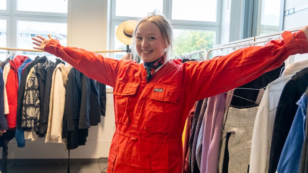 En person i rød jumpsuit står med utstrakte armer i en loppemerket, omgitt av klesstativer og et vindu i bakgrunnen.