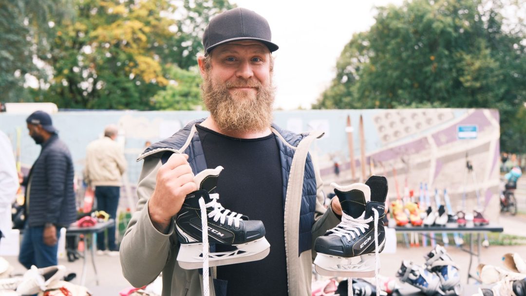 En skjeggete mann i caps holder stolt opp et par skøyter på et utendørs loppemarked, med travle mennesker og bord med eklektiske skatter i bakgrunnen.