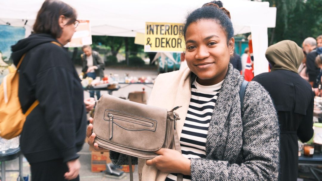 En person holder en grå veske ved en utendørs loppis, med folk som surfer i bakgrunnen.