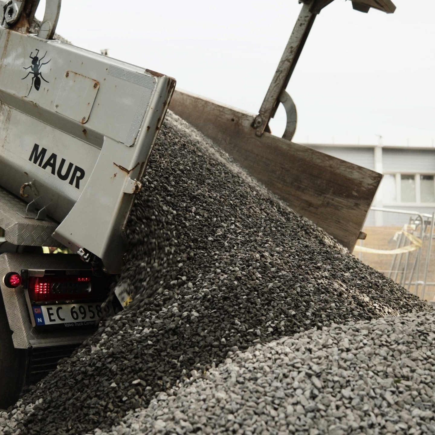På en travel byggeplass losser en dumper effektivt en grushaug, med forgangsutstyr og ruvende bygninger som står stolte i bakgrunnen.