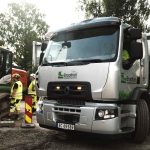 En hvit elektrisk lastebil står parkert på en byggeplass ved siden av arbeidere i verneutstyr. Trær og maskiner er i bakgrunnen.