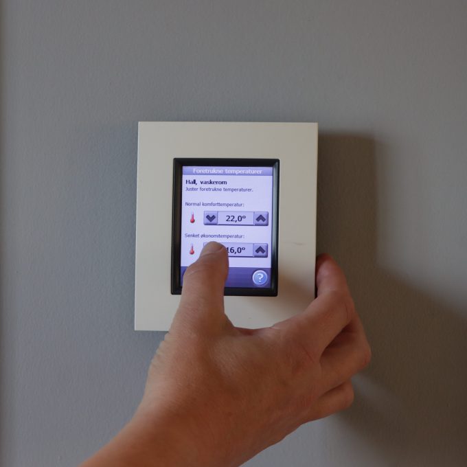 En hånd justerer temperaturen på en digital termostat montert på en grå vegg, og optimaliserer strømforbruket. Skjermen viser to temperaturinnstillinger, 22,0°C og 16,0°C, som inviterer deg til å spare energi effektivt.