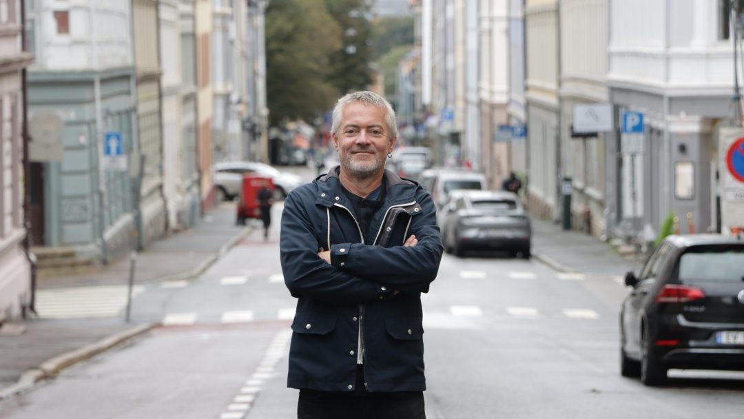 En mann med grått hår og skjegg står selvsikkert, med armene foldet, midt i en urban gate oppført på Byantikvarens Gul liste, omgitt av historiske bygninger og parkerte biler.