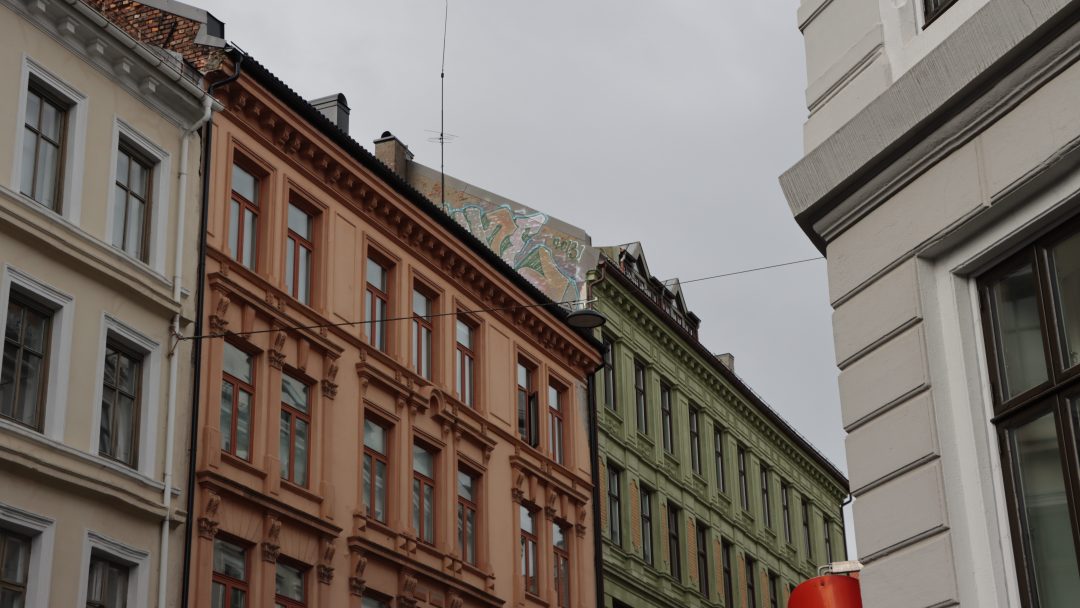 Rader med fargerike, historiske bygninger med utsmykkede detaljer står stolt mot en overskyet himmel. Et veggmaleri er delvis synlig på et hustak, og antyder Byantikvarens bevaringsarbeid for å vedlikeholde disse Gul liste-skattene.