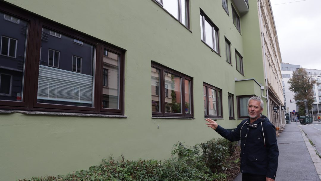 En person i mørk jakke gestikulerer mot vinduene i en grønn bygning i en bygate, nikker til dens historiske betydning, som Byantikvaren bemerket, og sørger for at den forblir på Gul-listen.