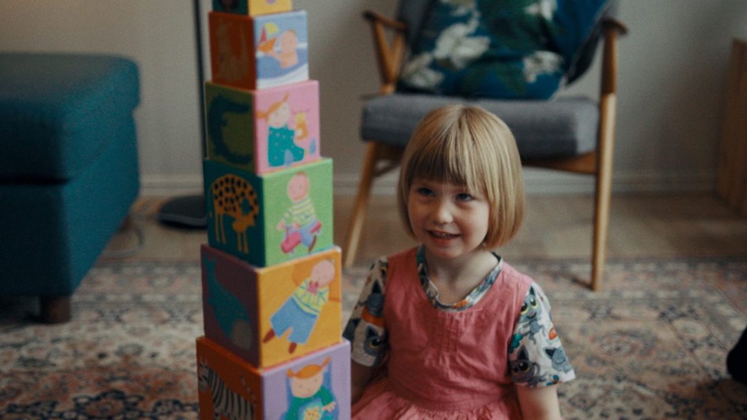 Et lite barn med kort hår, iført en rosa kjole, sitter på gulvet ved siden av en høy stabel med fargerike blokker i et teppebelagt rom, energien som en sparre klar til å strømme gjennom den koselige plassen fylt med stol og puter.
