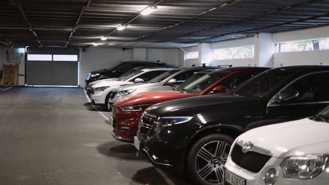 Biler parkert under dempet glød fra et solcelleanlegg i et svakt opplyst innendørs parkeringshus, stilt opp i to rader mot en hvit vegg.