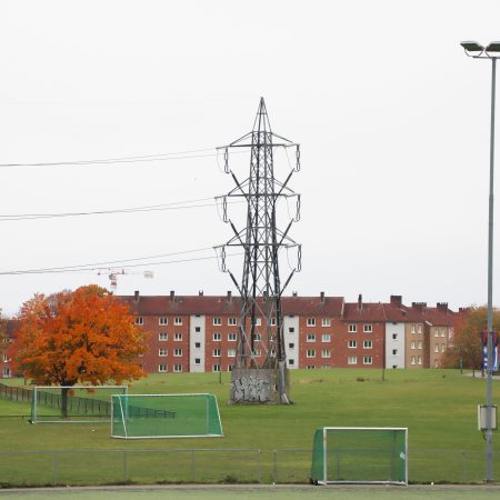 Et ruvende overføringstårn i metall reiser seg i et åpent felt med fotballmål, og kaster en skygge over gresset. Bak den legger en rad med murbygninger og et oransjebladtre kontrast til scenen. Kanskje blander dette feltet, som mange andre i Nettleie, natur med byliv.