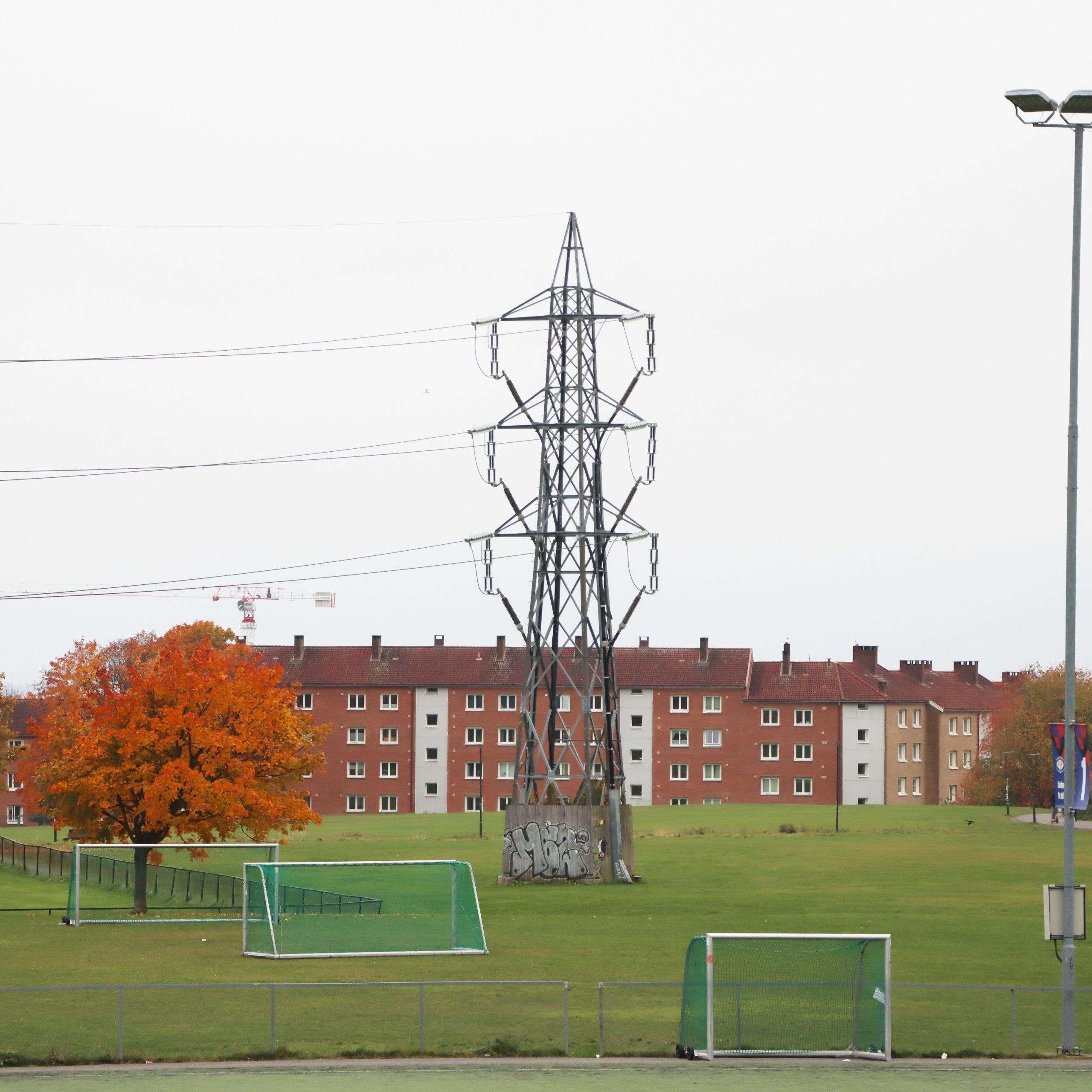 Et ruvende overføringstårn i metall reiser seg i et åpent felt med fotballmål, og kaster en skygge over gresset. Bak den legger en rad med murbygninger og et oransjebladtre kontrast til scenen. Kanskje blander dette feltet, som mange andre i Nettleie, natur med byliv.