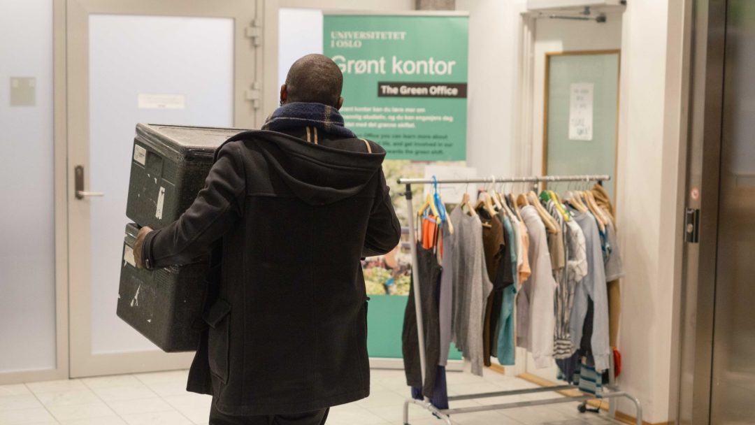En person bærer en stor boks inn i et rom. I nærheten står klær på stativ og møtemat-snacks er satt på bordet. På et skilt står det «Grønt kontor» ved Universitetet i Oslo.