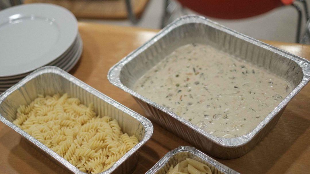 Aluminiumsbrett med kokt fusilli-pasta og kremet saus står på et trebord, klar til å tjene som perfekt møtemat, ved siden av en stabel med hvite tallerkener.
