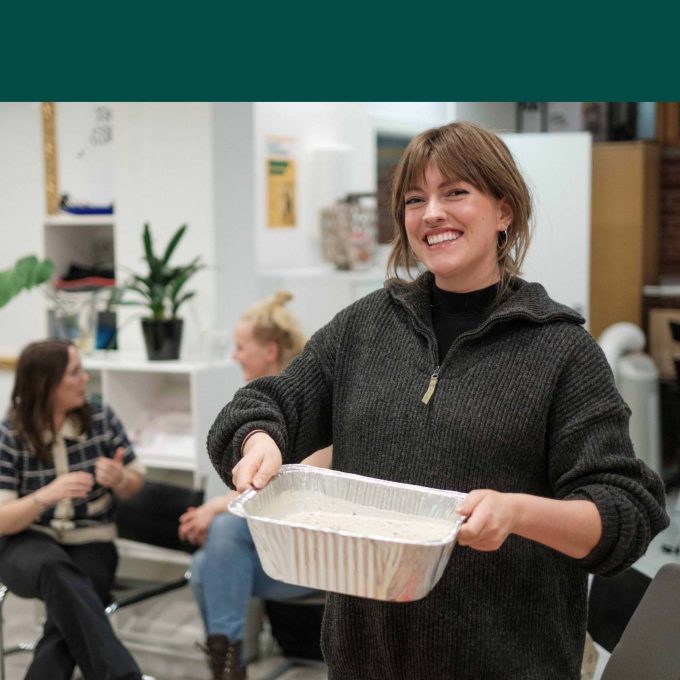 En person smiler, holder et metallbrett med møtematte, i et rom med folk og frodige planter i bakgrunnen.