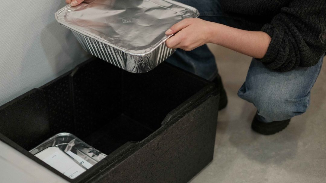 En person i jeans legger forsiktig møtemat, pent arrangert i en foliekledd beholder, i en isolert svart boks på gulvet.