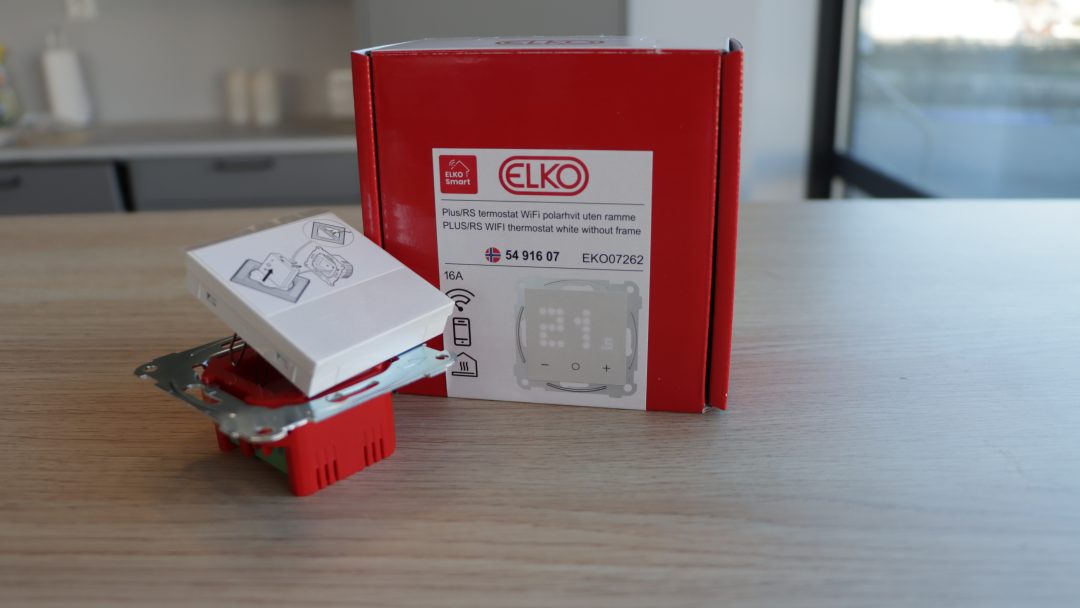 ELKO termostatboks og enhet, designet for smart strømstyring, sitter elegant på et trebord i et rom med solbelyst vindu.