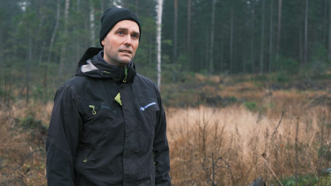 En person som har på seg en svart jakke og lue står i et skogkledd område og viser frem effekten av selektiv hogst, med tørt gress og selektivt tynnede trær i bakgrunnen.