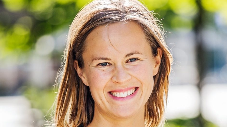 En person smiler utendørs, omgitt av den livlige uskarpheten av grønt løvverk, og tenker kanskje på strømsparetips mens de nyter naturens ro.