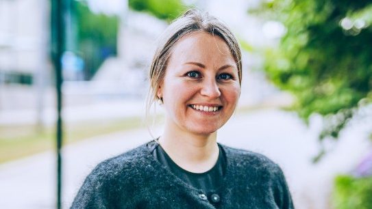 En smilende person med langt hår står utendørs i en grå genser, med uskarpt grønt i bakgrunnen, som minner om den frodige bevaringen som finnes i selektiv hogst-praksis.