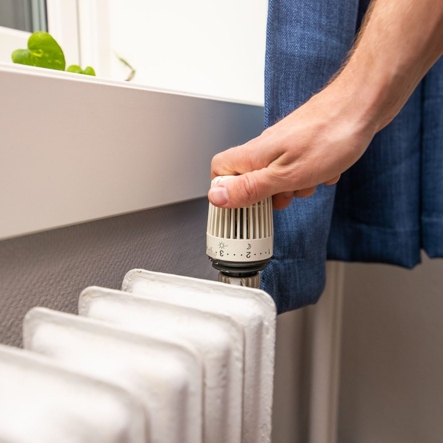 En hånd justerer forsiktig urskiven på en hvit radiator foran et vindu med en blå gardin, oppmerksom på spar strøm.