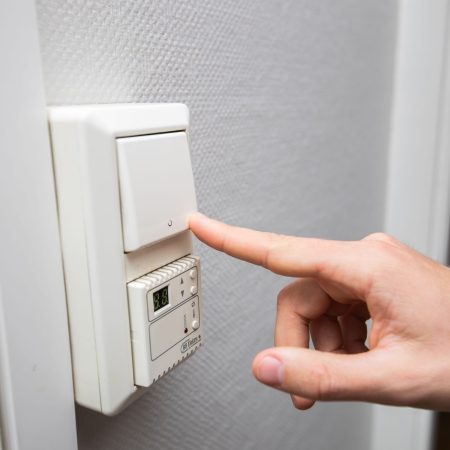En hånd strekker seg for å trykke på en lysbryter på veggen, og tilbyr strømsparetips, med et digitalt termostatdisplay under.