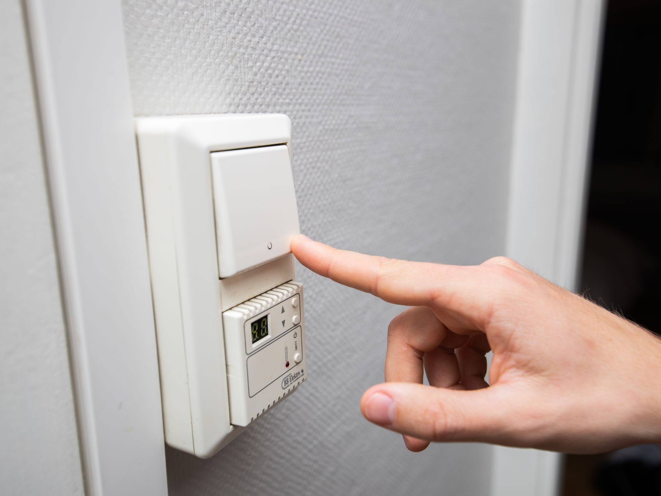 En hånd strekker seg for å trykke på en lysbryter på veggen, og tilbyr strømsparetips, med et digitalt termostatdisplay under.